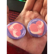 Köralakú kitűző lila alapon piros szívvel, két ölelő karral fehér Neurofibromatosis Warrior felirattal.