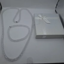 Átlátszó, csiszolt keskeny gyöngyökből álló nyaklánc és karkötő szett, fehér masnis díszdobozban.