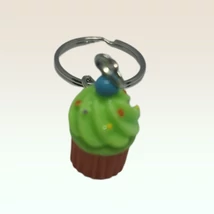 Ezüstszínű kulcskarikáról lánc lóg le, rajta medál: zöld habos muffin színes cukrokkal, kék gyümölccsel a tetején.