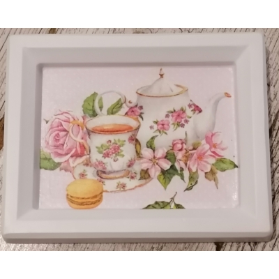 Fehér téglalapalakú gipsz képkeret közepén szalvéta kép: fehér, rózsaszín virágos kerámia teáscsésze és kancsó. Körülöttük rózsaszín virágok és egy