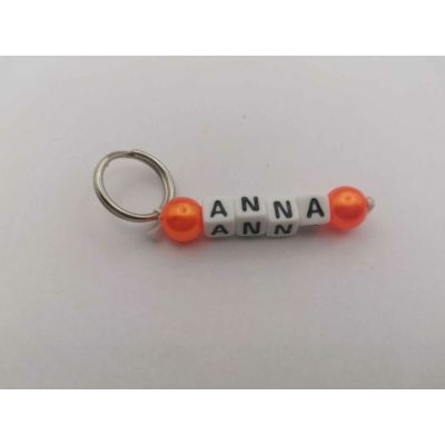 Ezüstszínű kulcskarikáról lelógó Anna szó betűgyöngyökből, két oldalán egy-egy gömbalakú, narancssárga gyönggyel. A betűgyöngyök kockaalakúak, fehér alapon feketék, elforgatva is ugyanazt mutatják.
