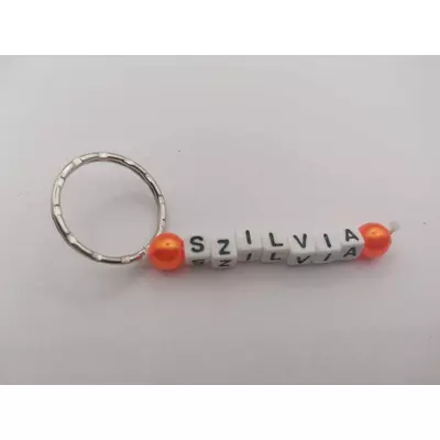 Ezüstszínű kulcskarikáról lelógó Szilvia szó betűgyöngyökből, két oldalán egy-egy gömbalakú, narancssárga gyönggyel. A betűgyöngyök kockaalakúak, fehér alapon feketék, elforgatva is ugyanazt mutatják. 