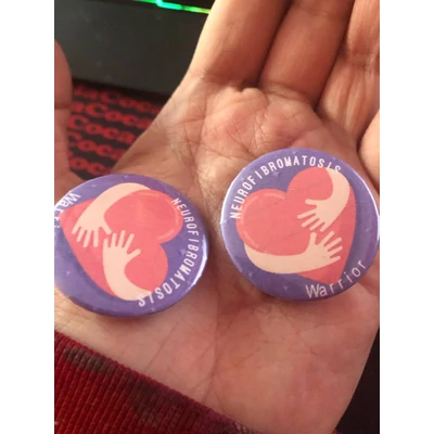 Köralakú kitűző lila alapon piros szívvel, két ölelő karral fehér Neurofibromatosis Warrior felirattal.