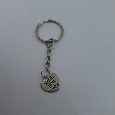 Ezüstszínű kulcstartó apró, szív alakú fém medállal, amelyen a „MADE FOR YOU” felirat olvasható. A medál egyszerű fém lánccal kapcsolódik a kulcskarikához.